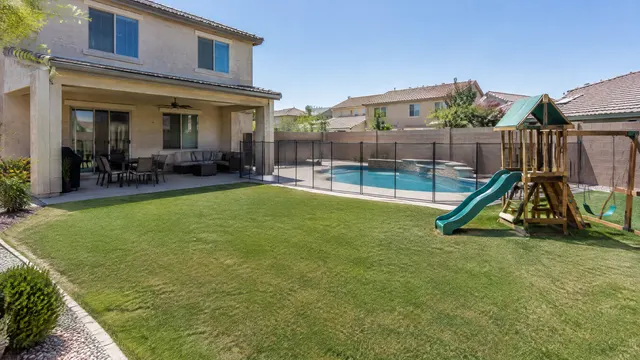 Backyard of a two-story family home in a Chandler, Arizona subdivision: artificial turf lawn with a small kids' play set, a pool fence, a covered patio and a block wall with neighboring roofs behind