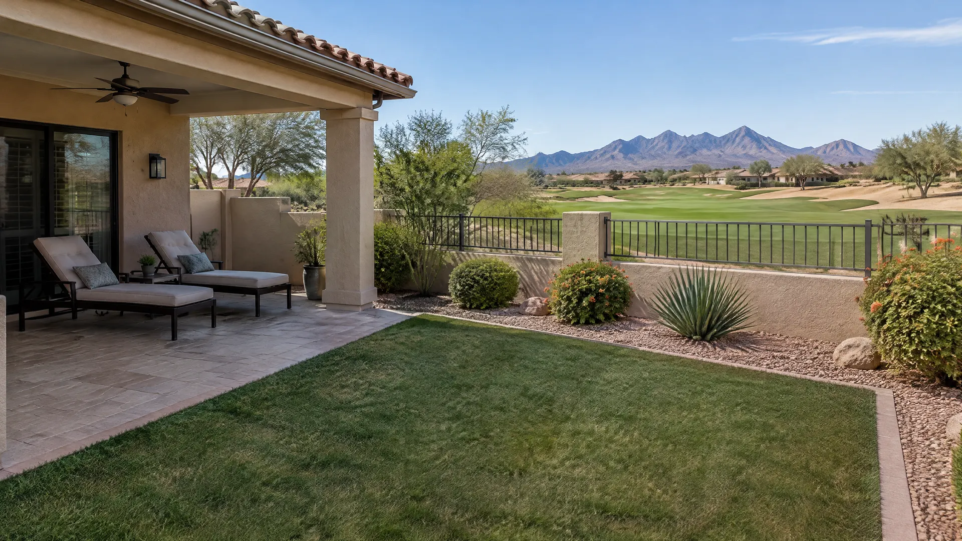 Backyard of a single-story Spanish-style home in a gated 55-plus community in south Chandler, Arizona: compact artificial turf lawn beside a covered patio with two lounge chairs, view to a golf course and the San Tan Mountains far away