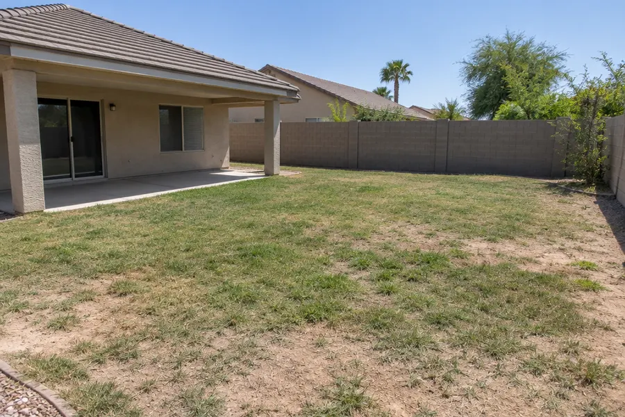 Backyard of a single-story Chandler, Arizona home before renovation: a worn, patchy, half-dead grass lawn with bare dirt spots, a simple concrete patio slab and a block wall, midday light