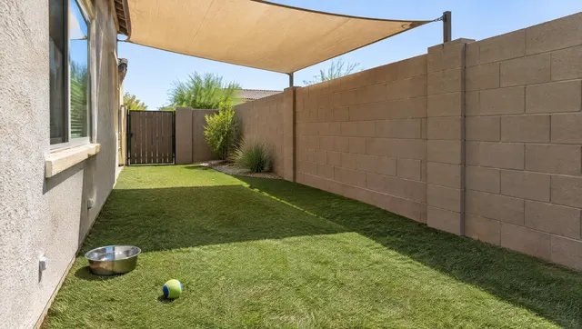 Side yard of a Chandler, Arizona home with durable pet turf: short dense artificial grass, a shade sail overhead, a water bowl and a dog ball on the turf, a block wall with a side gate, clean and tidy