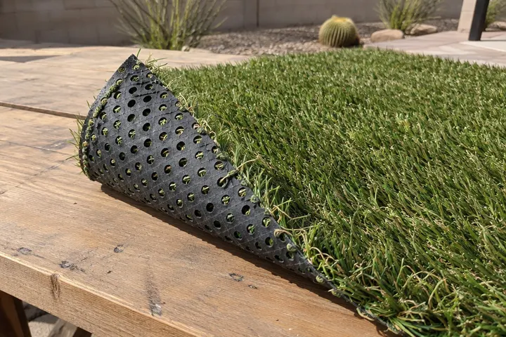 Corner of a piece of pet artificial turf flipped over to show its fully perforated flow-through backing, on a workbench